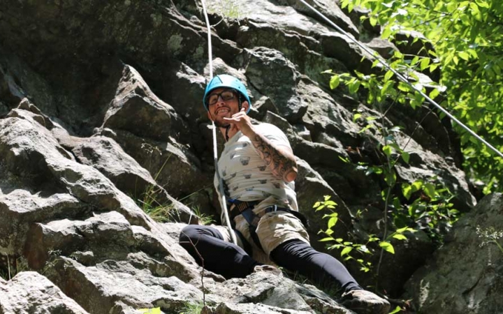 A person wearing safety gear and secured by ropes pauses a rock climbing exercise to give the camera a "hang loose" hand gesture. 